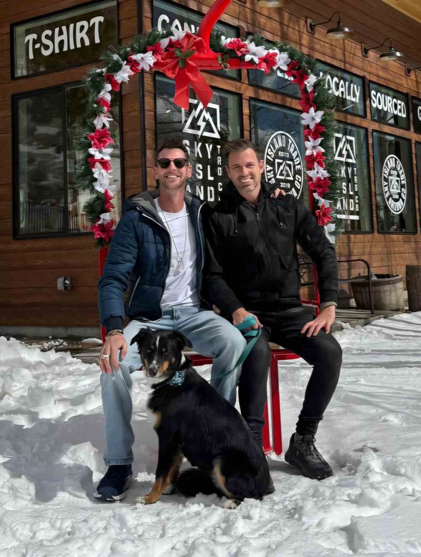 Kira, an Australian Shepherd, sitting on snow-covered ground on a leash between her two dads, all three smiling outside a wood-sided shop decorated with a holiday garland