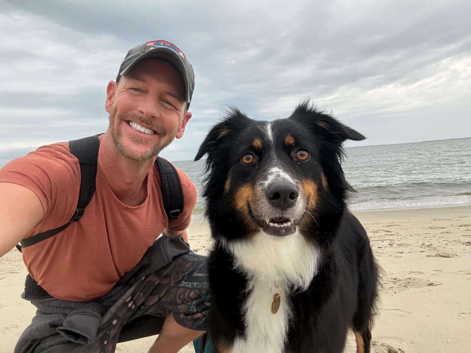 Kira sitting on a sandy beach beside one of her dads, both smiling for a selfie with the ocean behind them